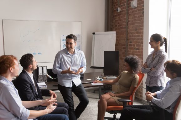 Multi-racial businesspeople gather at briefing, positive team leader talking to office employees interns or students at corporate training teach explain project strategy discuss business plan concept