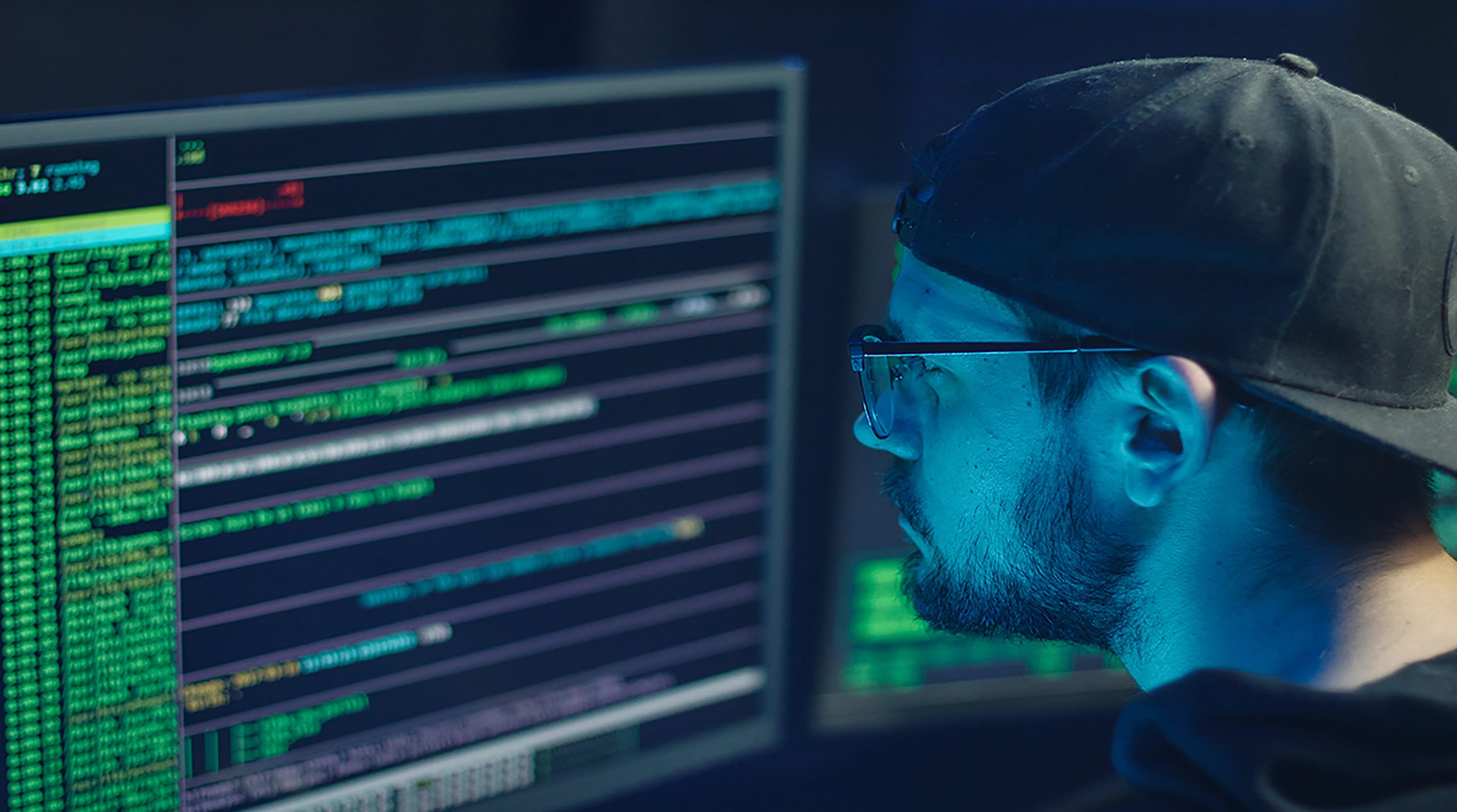 Man with a black cap and glasses looking at a computer screen with code on it - assumed to be a scammer or involved in some type of fraud